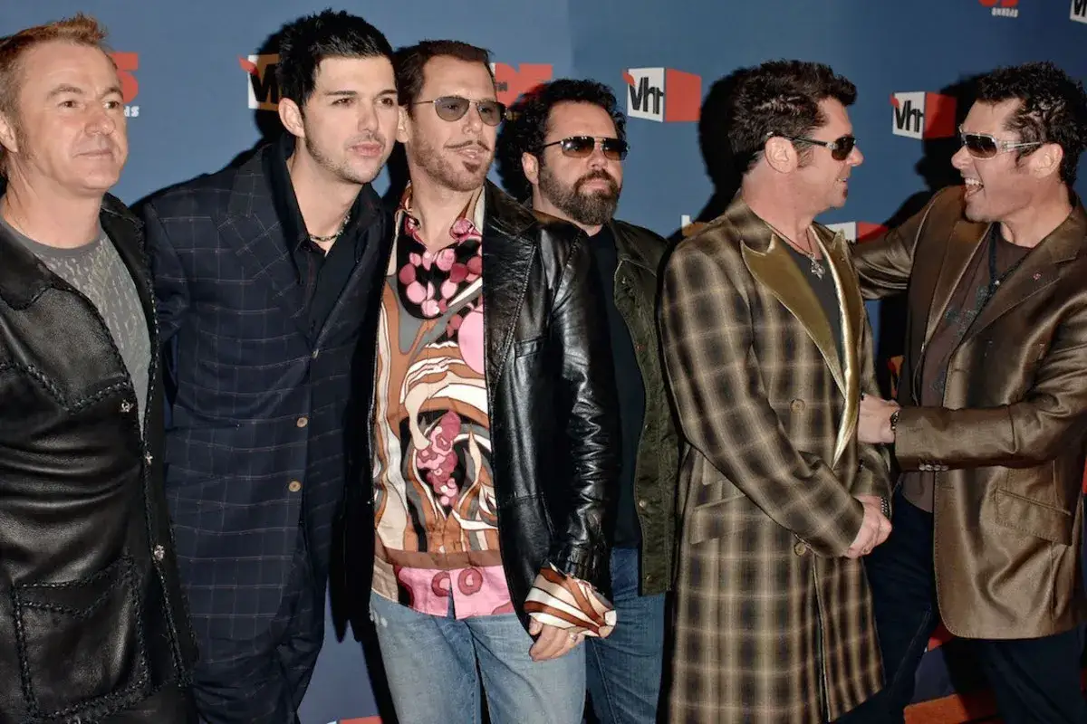 I membri degli INXS posano sorridenti, alcuni con occhiali da sole, su uno sfondo blu con il logo VH1.