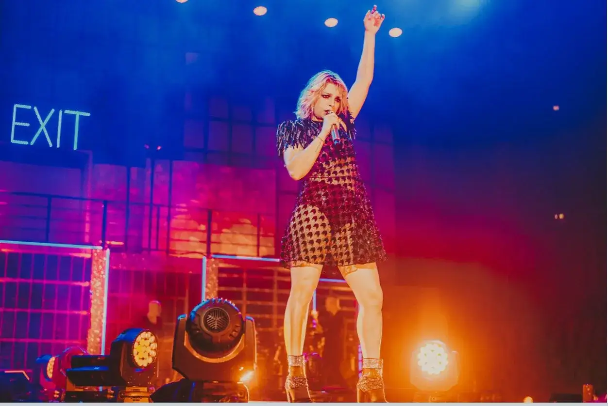 Emma Marrone canta sul palco, con un vestito scintillante e un braccio alzato. Le sue canzoni risuonano nell'aria, illuminate da luci intense.