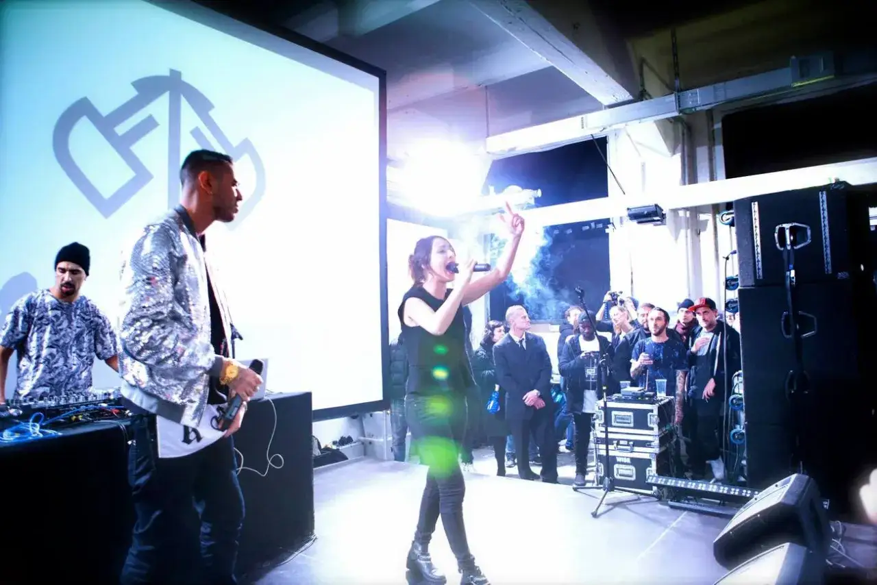 Federica Abbate canta sul palco, circondata da un pubblico attento. Un DJ mixa musica, mentre un altro artista con giacca scintillante la accompagna.