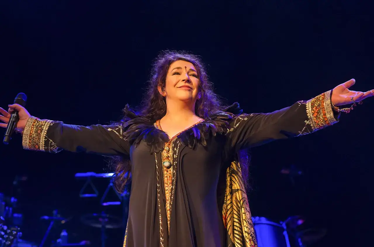 Una donna con un microfono, braccia aperte, evoca le canzoni di Kate Bush con un abito sontuoso e un'espressione sognante.