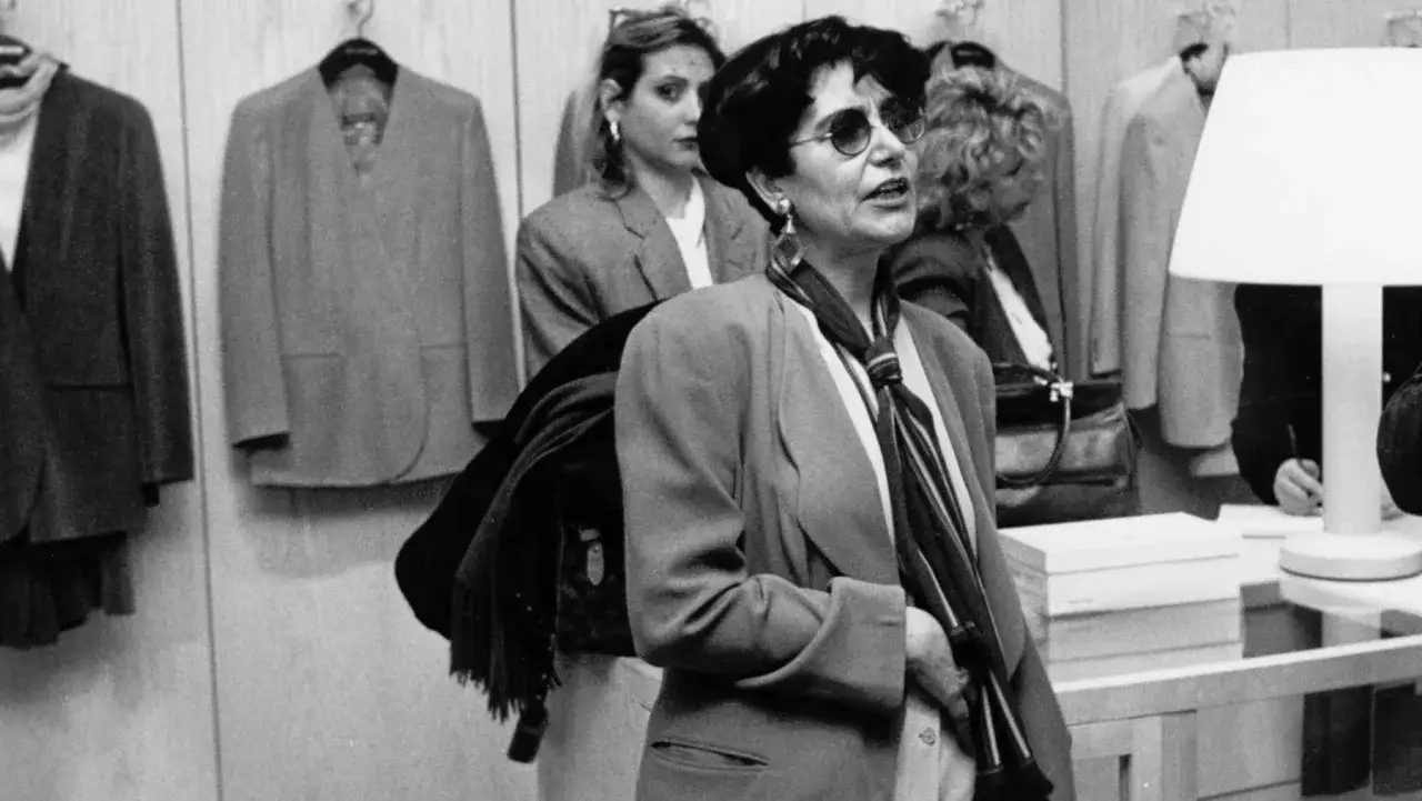 Una donna con occhiali da sole e una sciarpa a righe, forse Mia Martini, osserva capi d'abbigliamento in un negozio.