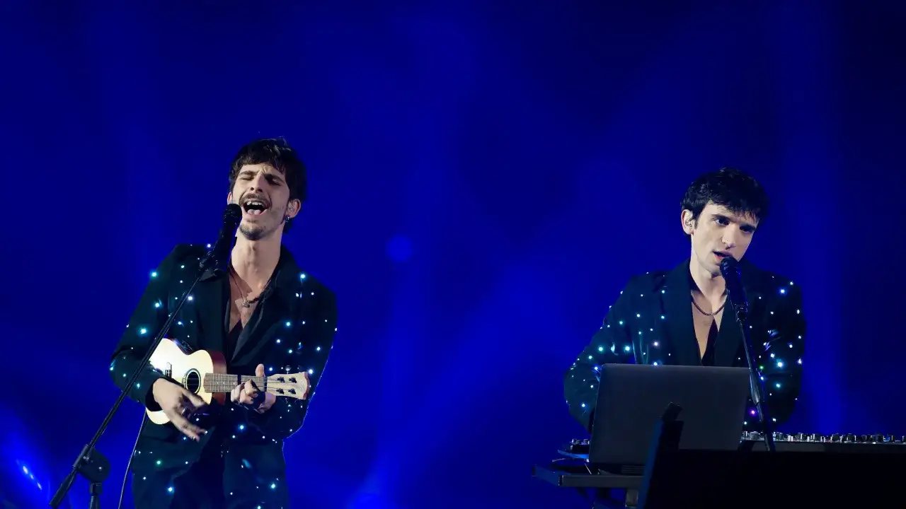 I Santi Francesi sul palco, uno canta con ukulele, l'altro alla tastiera, sotto un cielo blu stellato.