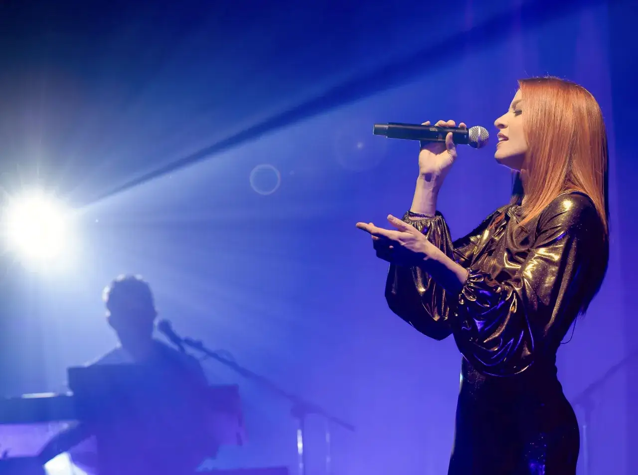 Noemi canta sul palco, illuminata da un fascio di luce. Le sue canzoni risuonano nell'aria, mentre un tastierista suona sullo sfondo.