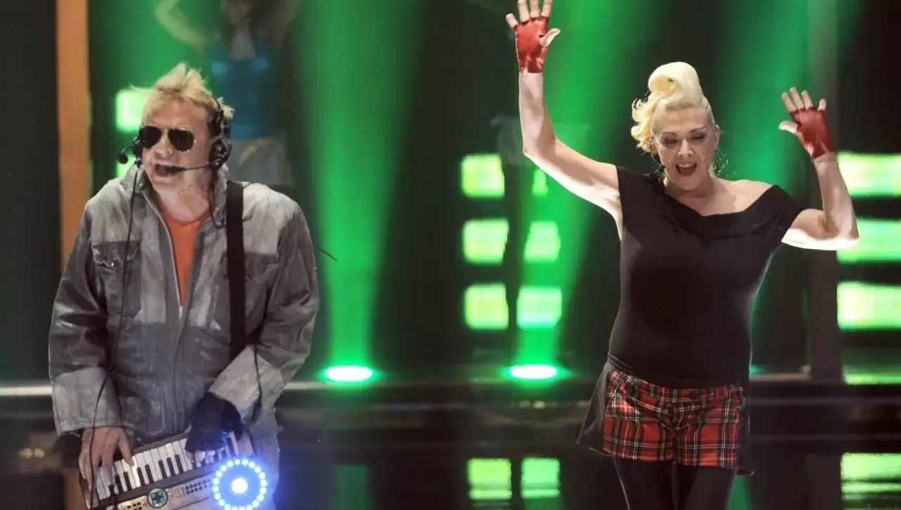 I membri dei Krisma, con look audaci, si esibiscono sul palco con luci verdi. Lui suona una tastiera, lei alza le mani.