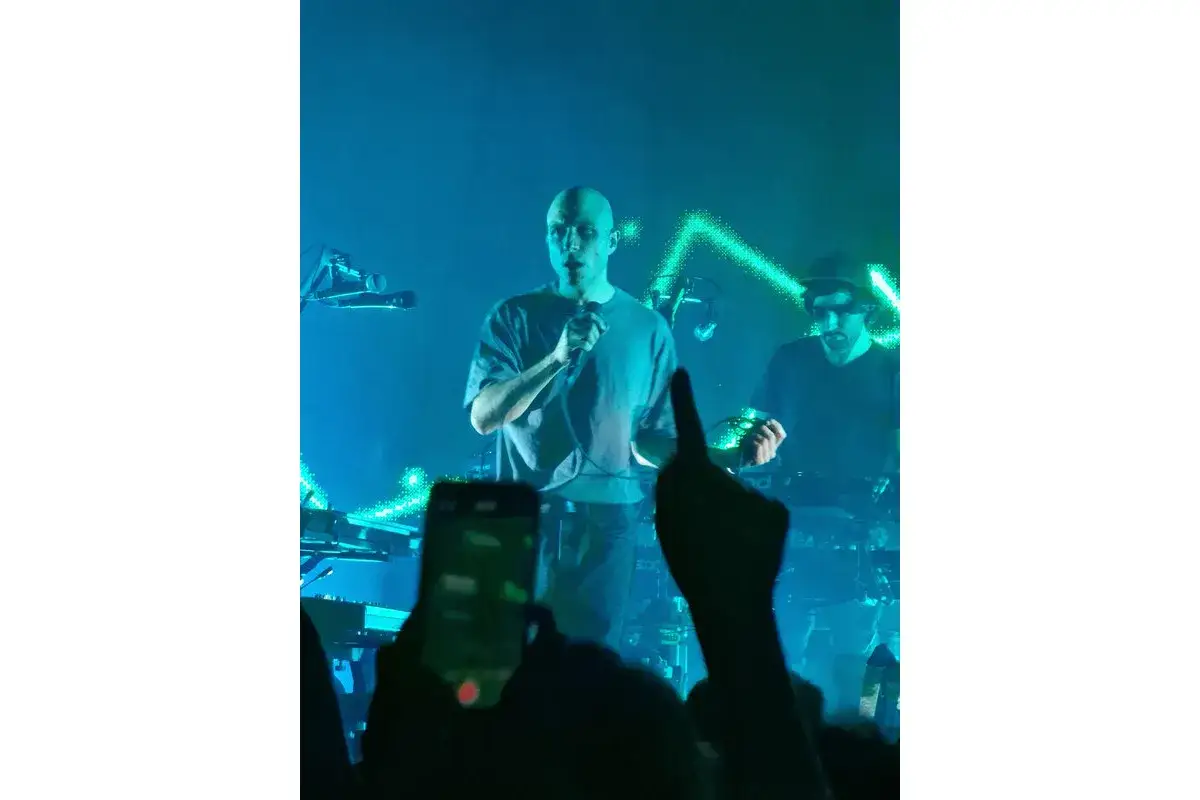 Sul palco, un uomo calvo canta al microfono, mentre un altro musicista suona dietro di lui. La scena &egrave; illuminata da luci blu e verdi, creando un'atmosfera da concerto. Sembra che la contessa e i cani stiano assistendo allo spettacolo.