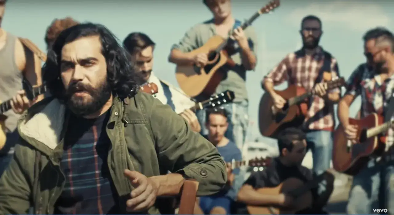 Un uomo con barba e capelli lunghi canta circondato da musicisti con chitarre, in un'atmosfera che ricorda 