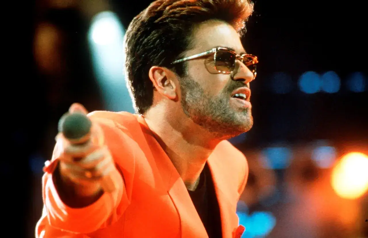George Michael cantante con occhiali da sole e giacca arancione, microfono in mano, sul palco.