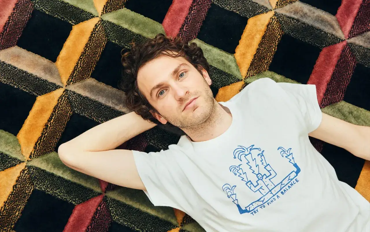 Un uomo con riccioli castani giace su un tappeto colorato, ispirato alle canzoni di Giorgio Poi. La sua maglietta bianca ha un disegno blu astratto.