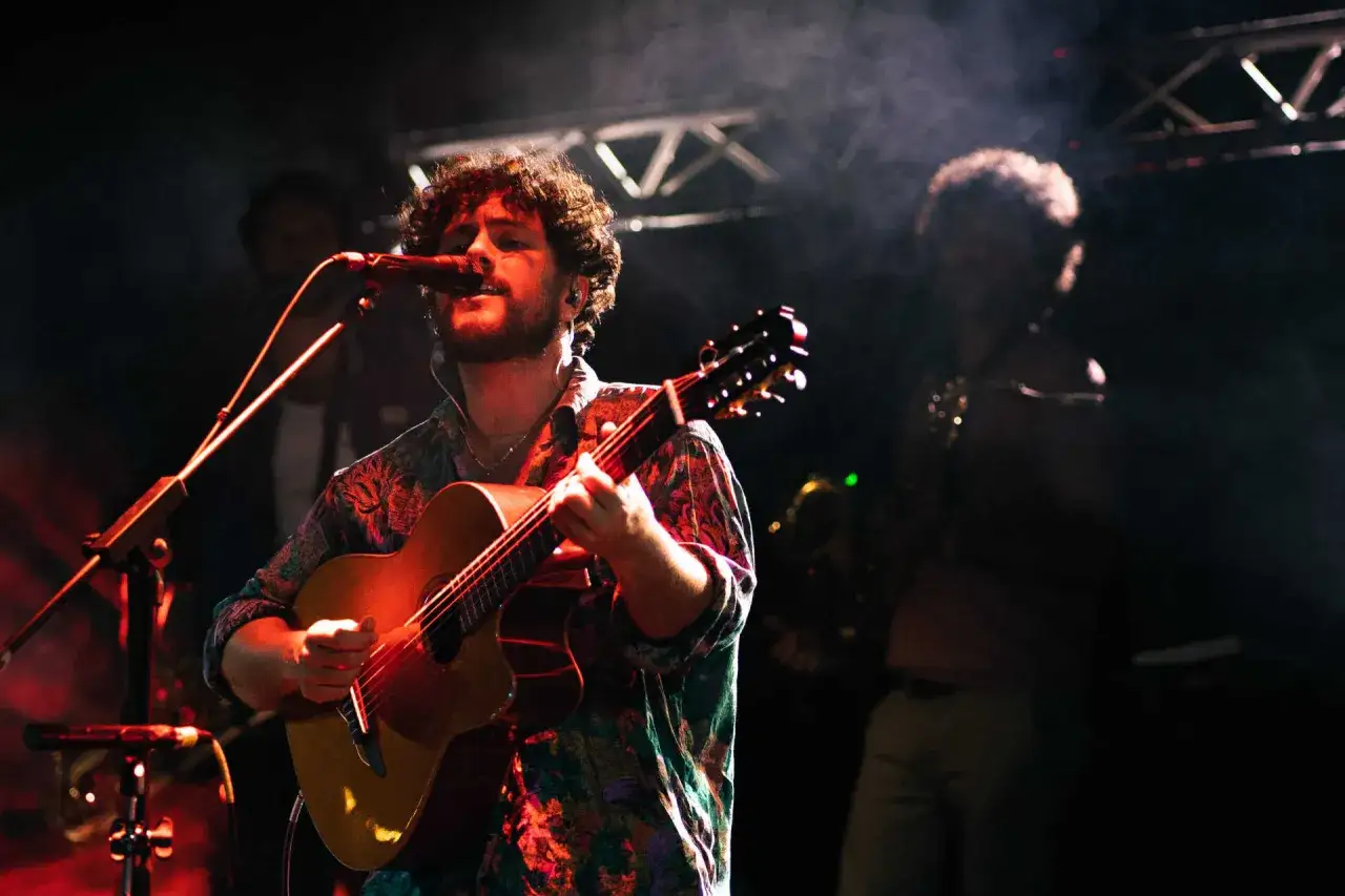 Marco Castello canta e suona la chitarra sul palco, illuminato da luci rosse e fumo.
