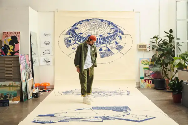 Un artista con un cappello arancione osserva disegni architettonici su carta, come se stesse creando nuove canzoni di Frah Quintale.
