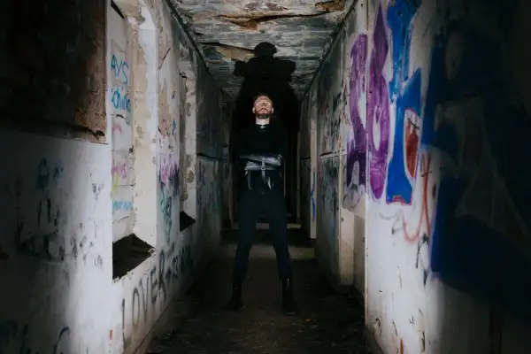 Uomo con barba in piedi in un corridoio pieno di graffiti, con indosso un abito nero. Sembra un rapper pronto per una sfida.
