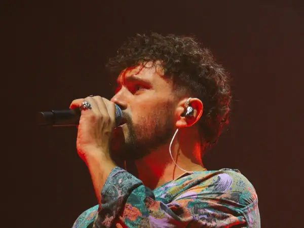 Marco Castello canta con passione, microfono in mano, cuffie nell'orecchio e un look colorato.