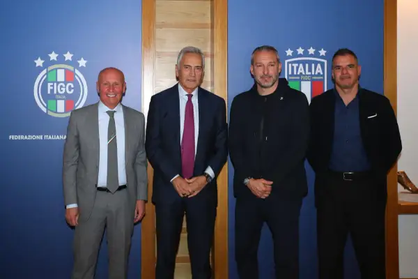 Quattro uomini in posa davanti a uno sfondo blu con il logo FIGC. Nessuno sembra un "dead can dance", ma tutti sono vestiti elegantemente.