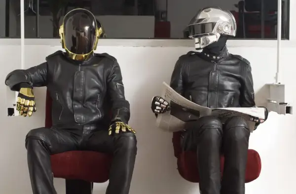 I Daft Punk, in pelle nera e caschi iconici, uno dorato e l'altro argentato, siedono leggendo il giornale.