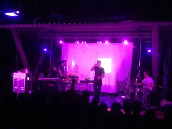 Concerto con luci viola. Sul palco, un cantante con occhiali e una tastiera. Dietro, un batterista e altri musicisti. Il pubblico è in ombra.