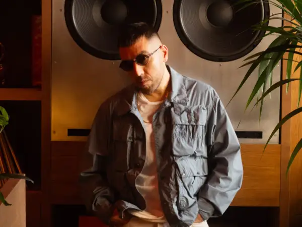 Man in sunglasses and a grey jacket leans against a large speaker.