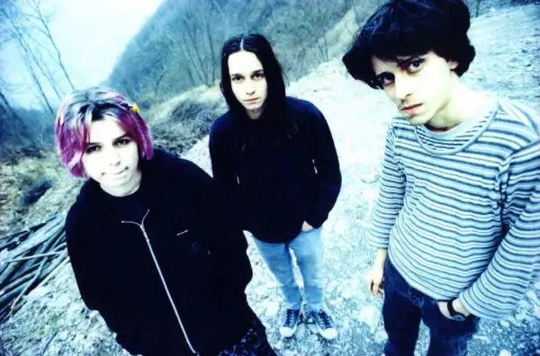 Three young people with dark hair and casual clothing stand outdoors in a rocky, wooded area.