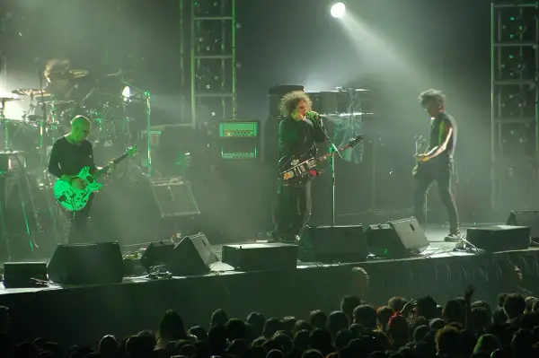 Sul palco, i membri dei The Cure suonano le loro iconiche canzoni. Un chitarrista con una chitarra verde brillante, il cantante con la sua chitarra nera e un altro chitarrista.