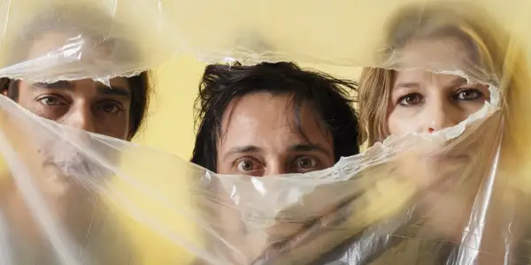 Occhi curiosi di Alberto Ferrari Verdena e altri due volti emergono da un telo di plastica trasparente su sfondo giallo.