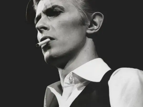 David Bowie, un'icona dei suoi golden years, produttore di musica e stile, fuma una sigaretta con sguardo intenso.