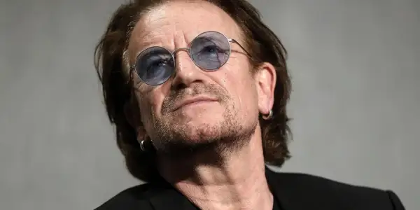 Primo piano di Bono Vox con occhiali rotondi blu, che ricorda le sue iconiche canzoni.