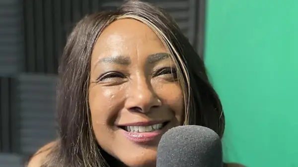 Dee C. Lee sorride vicino a un microfono, con uno sfondo verde e pannelli acustici.
