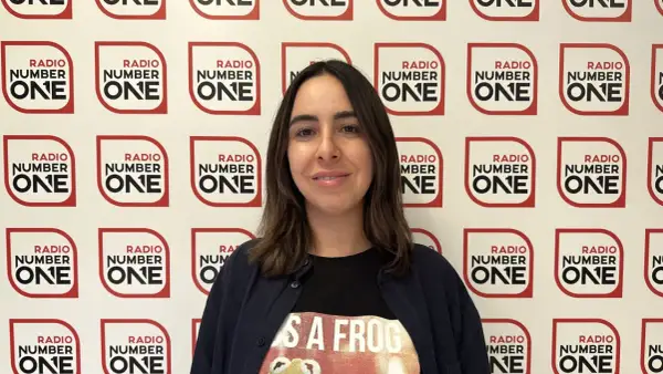 Federica Abbate, con una maglietta a tema rana, sorride davanti a uno sfondo con il logo di Radio Number One. Le sue canzoni risuonano in radio.