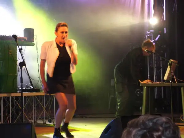 Sul palco, i membri di Krisma cantano e suonano. Una donna con giacca bianca e gonna nera è in primo piano, mentre un uomo è dietro un mixer.