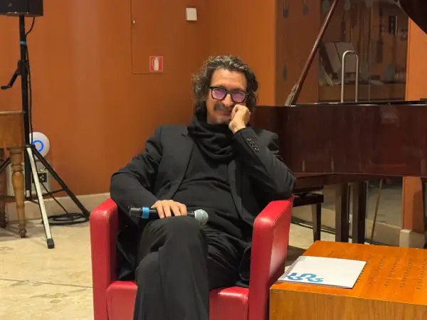 Sergio Cammariere, con microfono in mano, siede su una poltrona rossa, pronto a raccontare le sue canzoni.