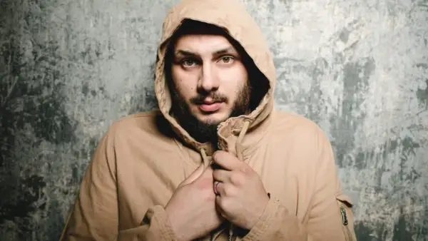 Uomo con cappuccio beige, sguardo intenso, barba. Sembra che qualcuno abbia sbagliato con lui.